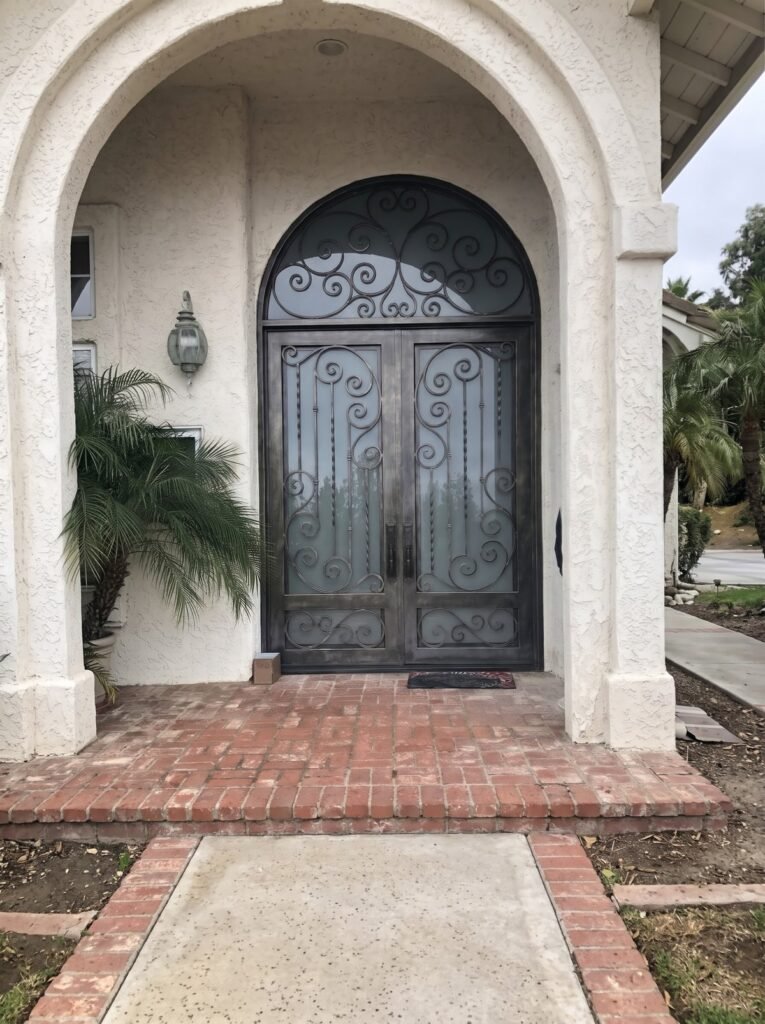 Arched wrought iron double entry door with scrollwork and frosted glass under a stucco archway with brick porch