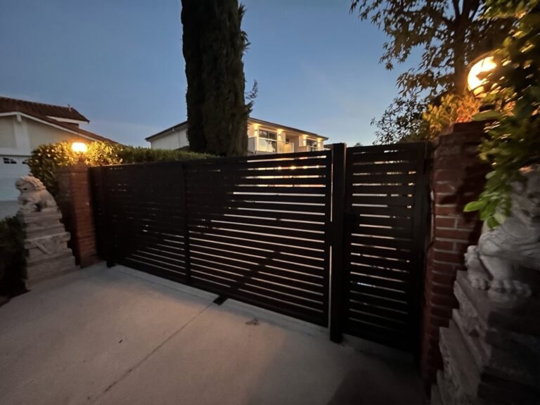 Modern black horizontal slat steel driveway gate and matching pedestrian gate between brick pillars at dusk